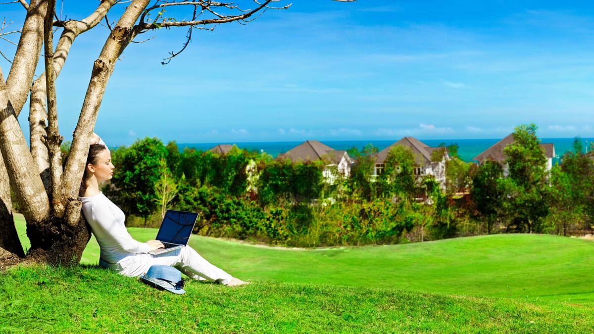Pretty woman sitting with laptop under tree. Beautiful panoramic view of idyllic landscape